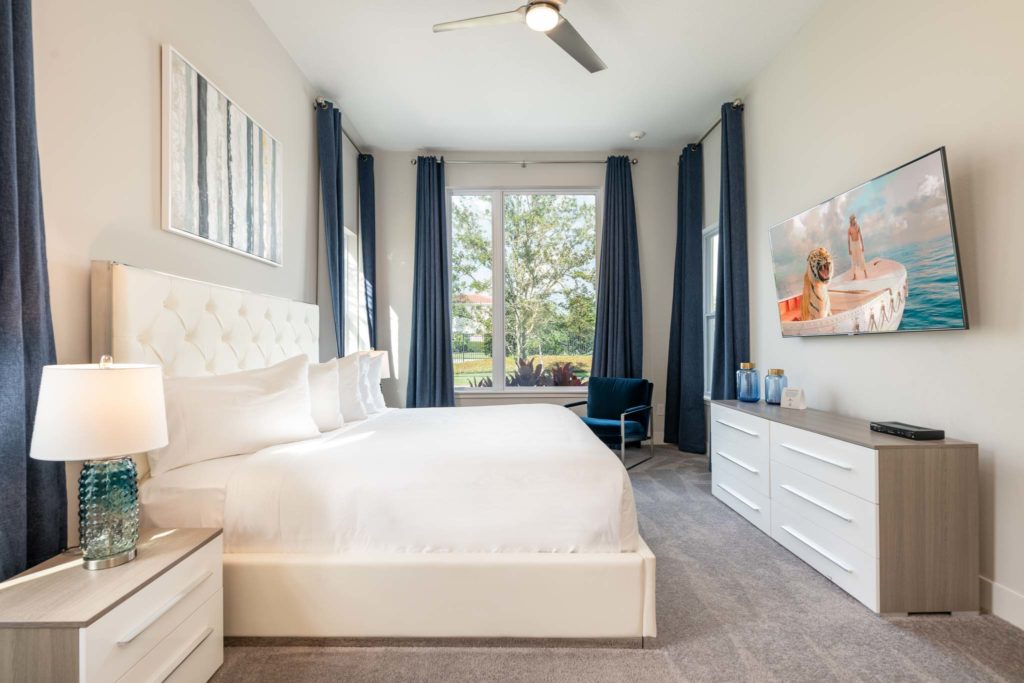 Bedroom with large king bed, plush headboard, two side tables, armchair, wall-mounted TV, long dresser, and four large open windows.