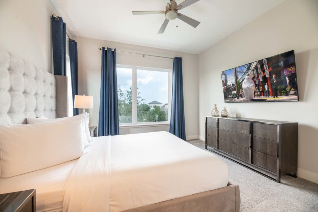 Bedroom with large king bed, plush headboard, wall-mounted TV, ceiling fan, long dresser, and two large open windows.