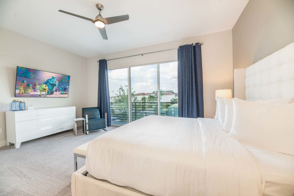 Spacious bedroom with large king bed, plush headboard, wall-mounted TV, long dresser, and sliding glass door balcony access.