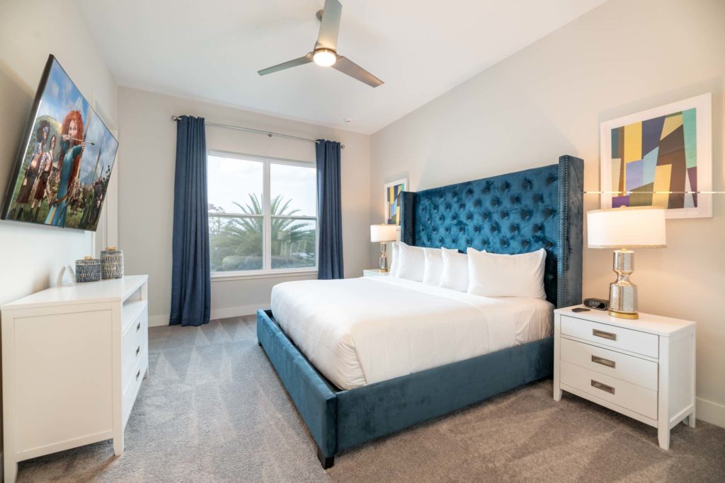 Bedroom with large king bed, plush suede headboard, wall-mounted TV, long dresser, ceiling fan, side tables, and large window.