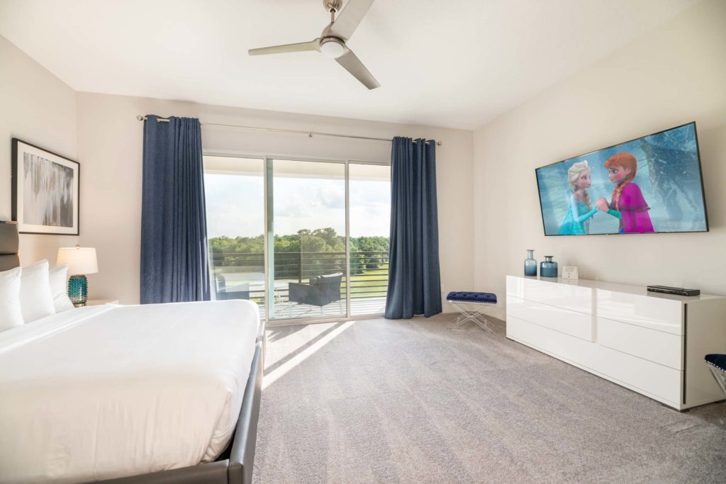 Spacious bedroom with large king bed, leather headboard, wall-mounted TV, long dresser, and sliding door balcony access.