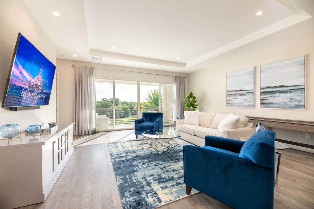 Upper loft with shuffleboard table, sofa, armchairs, wall-mounted TV, and sliding door balcony access.