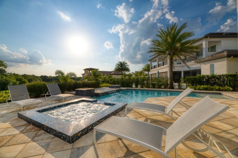 Private backyard patio with pool, hot tub, waterfall, fire pits, and lounge chairs on a sunny afternoon.