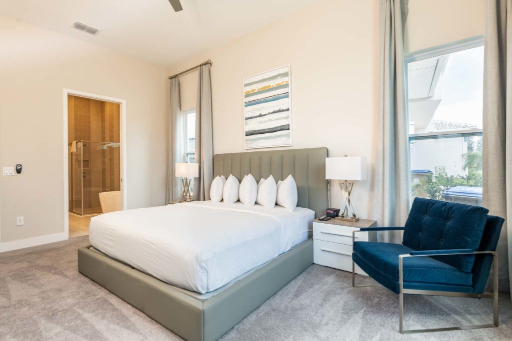 Bedroom with large king bed, plush headboard, modern armchair, decorative wall art, door to bathroom, and large window.