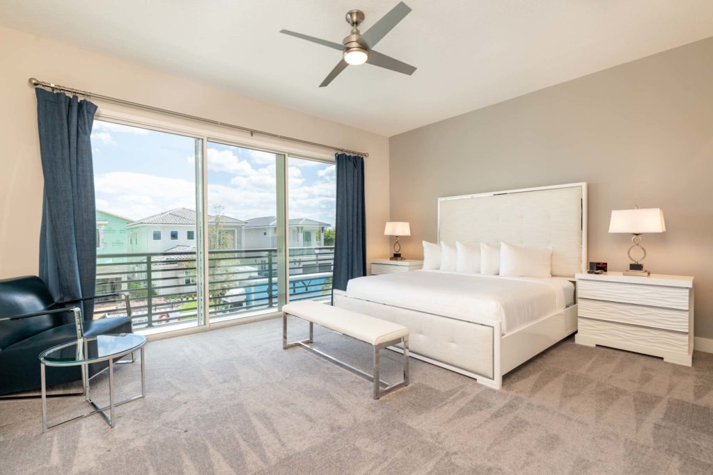 Spacious bedroom with large king bed, plush headboard, ceiling fan, modern armchair, and sliding door access to balcony.