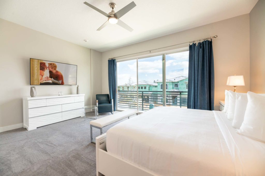 Spacious bedroom with large king bed, wall-mounted TV, armchair, glass-surface side table, and sliding door balcony access.