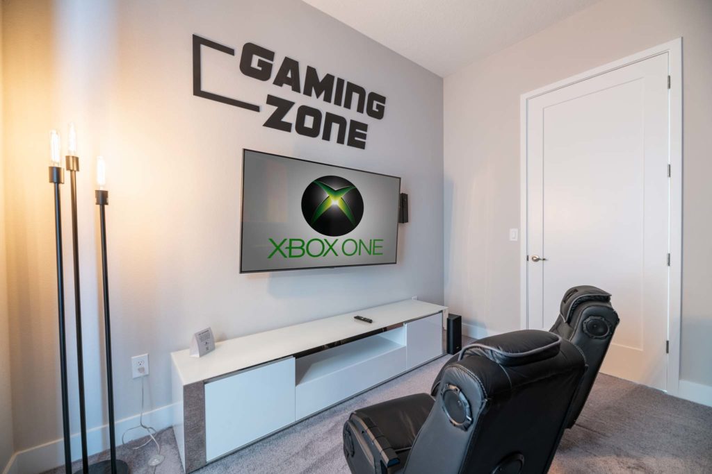 Game room with “Gaming Zone” wall mounted sign, TV playing Xbox One, and two rocking gaming chairs.