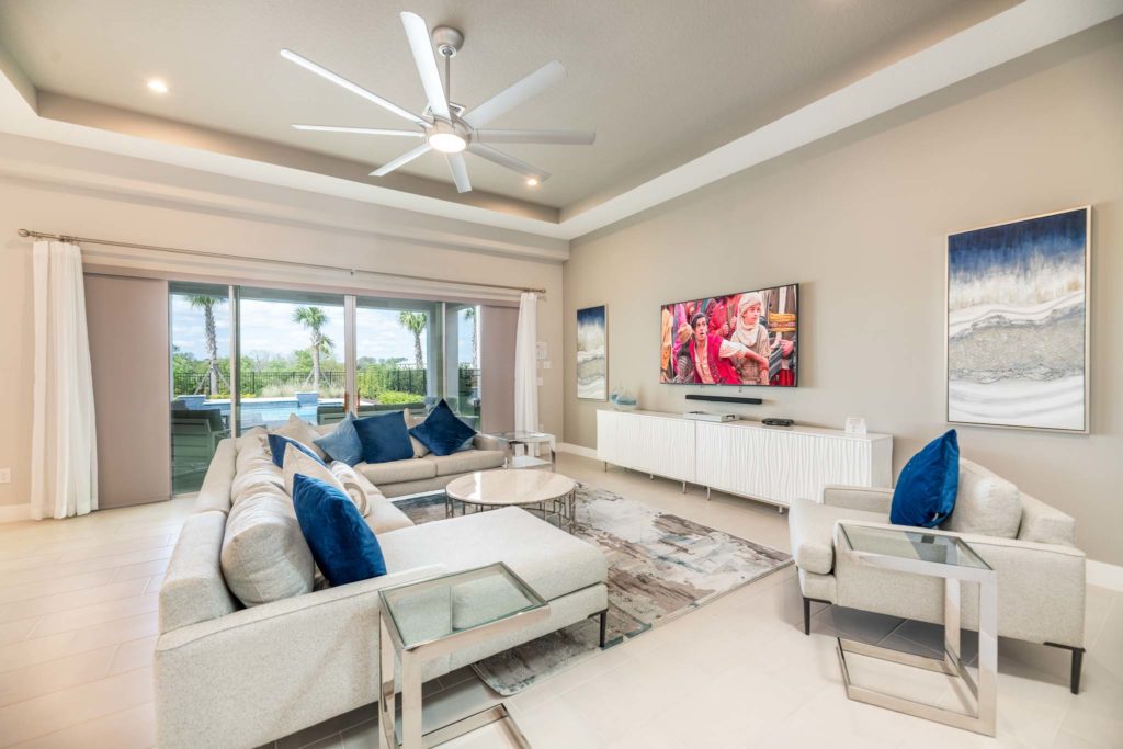 Spacious living room with sectional sofa, wall-mounted TV, decorative wall art, large ceiling fan, and sliding door access to private backyard pool.